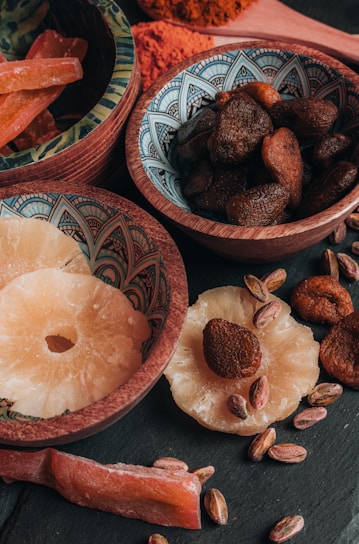 An assortment of dried fruits and nuts is arranged in decorative bowls and scattered on a dark surface. The bowls contain dried apricots, pistachios, and slices of dried pineapple. The intricate patterns on the bowls add an artistic touch to the rustic presentation.
