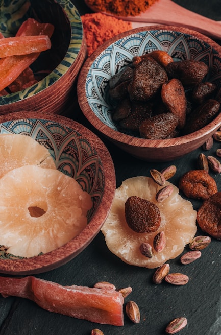 An assortment of dried fruits and nuts is arranged in decorative bowls and scattered on a dark surface. The bowls contain dried apricots, pistachios, and slices of dried pineapple. The intricate patterns on the bowls add an artistic touch to the rustic presentation.