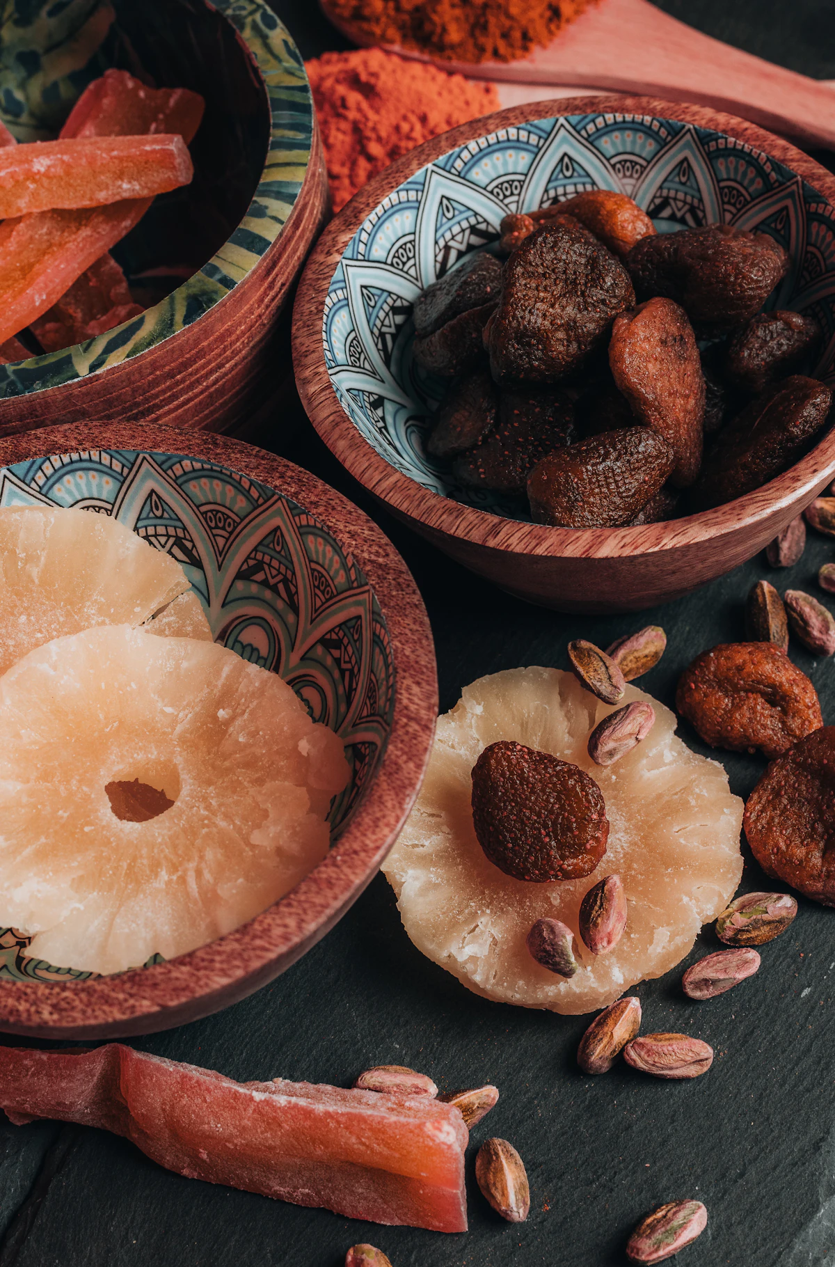 Dried pineapple, dried figs, and pistachios in patterned bowls on slate
