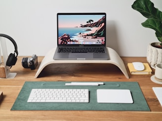 A minimalist desk setup featuring a laptop on a wooden stand, displaying a colorful digital landscape wallpaper. Surrounding the laptop are various items including headphones on a stand, a vintage camera, a keyboard, a trackpad, a pen, a notepad with a mouse, and a potted plant. The wooden desk and accessories contribute to the neat and organized aesthetic.