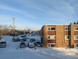 A snowy residential area with cars parked along the street and in a parking lot. The brick apartment building has several windows, and snow covers the ground and the vehicles. Power lines are visible above, and a few trees and a fence can be seen in the background.