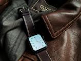 A sophisticated watch with a square face and a brown leather strap is resting on a textured surface made of brown leather and dark fabric. Visible stitching on the leather gives a sense of craftsmanship and quality.