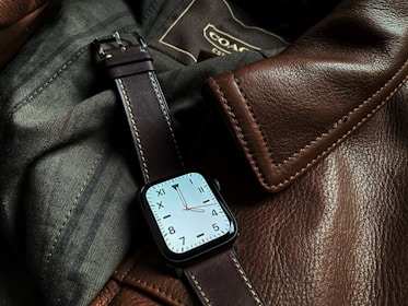 A sophisticated watch with a square face and a brown leather strap is resting on a textured surface made of brown leather and dark fabric. Visible stitching on the leather gives a sense of craftsmanship and quality.