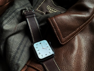 A sophisticated watch with a square face and a brown leather strap is resting on a textured surface made of brown leather and dark fabric. Visible stitching on the leather gives a sense of craftsmanship and quality.