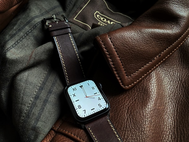 A sophisticated watch with a square face and a brown leather strap is resting on a textured surface made of brown leather and dark fabric. Visible stitching on the leather gives a sense of craftsmanship and quality.