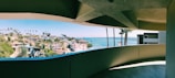 A panoramic view of a coastal city seen through a curved, shaded balcony. The cityscape features numerous modern houses surrounded by lush greenery and tall palm trees. Beyond the city lies a calm, blue ocean extending to the horizon under a clear sky.