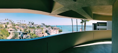A panoramic view of a coastal city seen through a curved, shaded balcony. The cityscape features numerous modern houses surrounded by lush greenery and tall palm trees. Beyond the city lies a calm, blue ocean extending to the horizon under a clear sky.