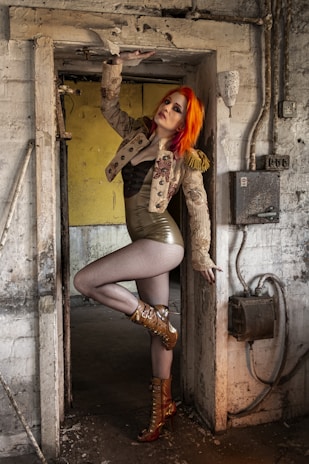 A person with vibrant red-orange hair is posing in a doorway of an old, industrial setting. They are wearing a decorated jacket with tassels, a shiny form-fitting outfit, fishnet stockings, and high boots with intricate designs. The walls are textured and worn, with exposed pipes and a yellow background visible through the door.