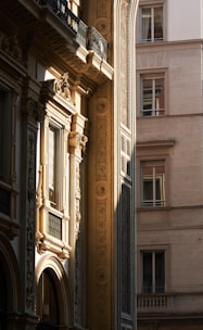 Intricately designed architectural facade with classical elements such as ornate window frames, detailed carvings, and decorative motifs. The image captures the warm sunlight casting shadows on this historic building, highlighting its detailed craftsmanship. Adjacent is a modern building with simpler design, contrasting the architectural styles.