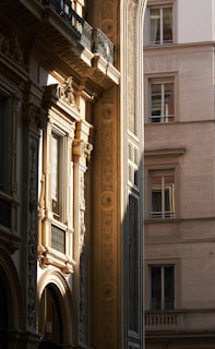 Intricately designed architectural facade with classical elements such as ornate window frames, detailed carvings, and decorative motifs. The image captures the warm sunlight casting shadows on this historic building, highlighting its detailed craftsmanship. Adjacent is a modern building with simpler design, contrasting the architectural styles.