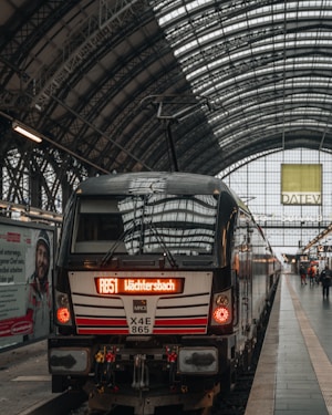 A modern train is stationed at a bustling railway platform under a large arched glass and metal roof. The platform is populated by a few people, and there is a prominent advertisement on the platform wall. The destination board on the train reads 'W&auml;chtersbach' with the route number 'RB51'. The train tracks and the platform are clean and well-maintained.