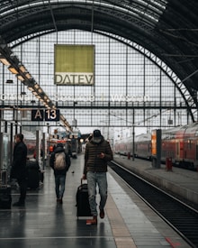 A train station platform with people walking and waiting. One person is pulling a suitcase and looking at their phone. The platform is covered by an arched glass ceiling with visible signage and advertisements. Trains are parked on the tracks beside the platform.