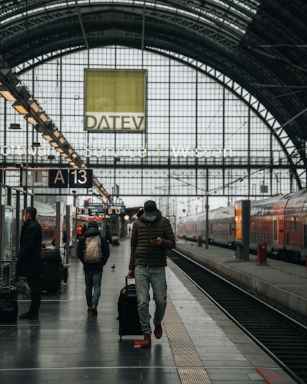 A train station platform with people walking and waiting. One person is pulling a suitcase and looking at their phone. The platform is covered by an arched glass ceiling with visible signage and advertisements. Trains are parked on the tracks beside the platform.