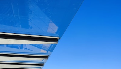 A modern architectural detail featuring horizontal slats or beams beneath a clear or lightly tinted blue glass structure, set against a bright blue sky.