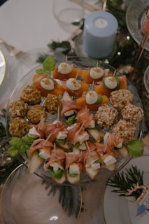 An elegant arrangement of assorted appetizers on a glass platter includes skewers with cheese, cured meat, and vegetables. Savory balls with coating and garnished skewers are visible. A blue candle and greenery add to the decorative background.