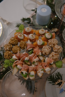 An elegant arrangement of assorted appetizers on a glass platter includes skewers with cheese, cured meat, and vegetables. Savory balls with coating and garnished skewers are visible. A blue candle and greenery add to the decorative background.
