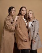 Three women stand closely together against a beige background, each wearing a different shade of trench coat. The woman on the left has dark hair and her hand gently rests on the shoulder of the woman in the middle, who has medium-length brown hair. The woman on the right has blond hair and is looking forward with her hands folded in front.