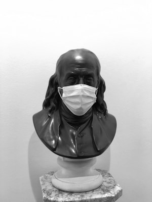 A black and white bust sculpture is wearing a surgical mask. The sculpture is positioned on a marble pedestal against a plain wall backdrop. The bust has long, flowing hair and is dressed in period attire.