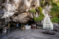 A natural grotto featuring rugged stone surfaces and greenery. A statue is positioned in a niche, surrounded by lush vegetation. Several people are standing near the statue, observing and possibly praying. A tall metal holder filled with numerous lit white candles is present, adding a spiritual element.