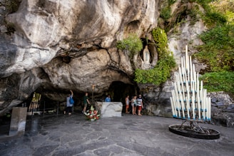 A natural grotto featuring rugged stone surfaces and greenery. A statue is positioned in a niche, surrounded by lush vegetation. Several people are standing near the statue, observing and possibly praying. A tall metal holder filled with numerous lit white candles is present, adding a spiritual element.