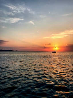 The image captures a serene sunset over the ocean, with a sailboat silhouetted against the vibrant orange and pink hues of the sky. The water reflects the colorful sky, creating a shimmering effect. Gentle waves create texture on the ocean's surface, while clouds add depth to the scene.