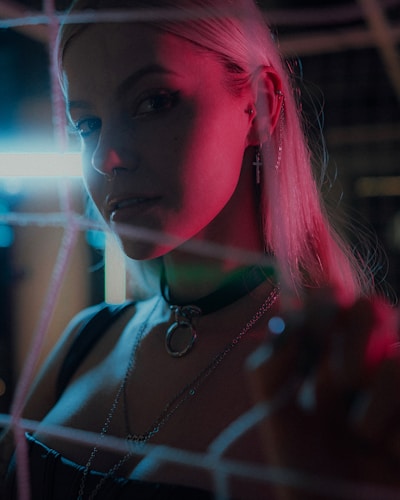 A person with long hair is illuminated by colorful lighting, featuring hues of pink and blue. They wear multiple ear piercings, including a cross earring, and a choker necklace with a large ring. The background appears urban with hints of neon lighting.