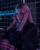 A person with long blonde hair and a nose ring is positioned against a neon-lit grid background. They are holding their head with both hands, wearing a black jacket and patterned tights. The lighting creates a dramatic contrast with purples and blues.