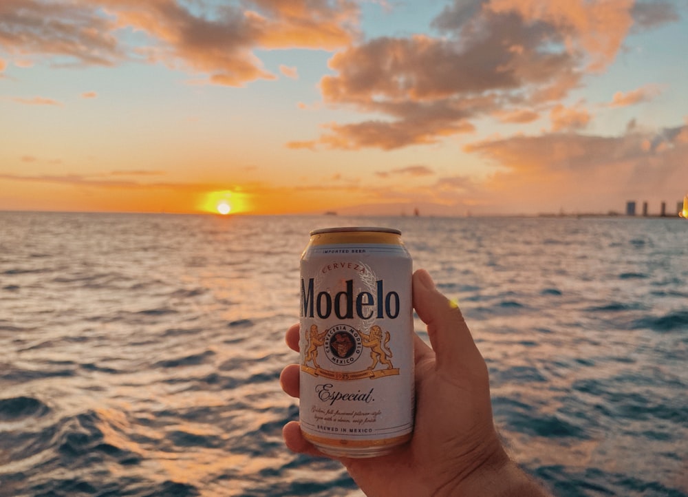 a hand holding a can of beer in front of the ocean