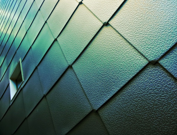 A wall covered with textured, overlapping metal panels in a teal hue, featuring a small rectangular window. The panels are arranged in a diagonal pattern, creating a sense of depth and modernity.