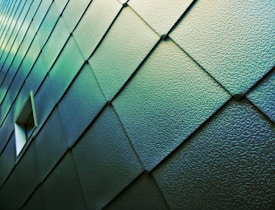 A wall covered with textured, overlapping metal panels in a teal hue, featuring a small rectangular window. The panels are arranged in a diagonal pattern, creating a sense of depth and modernity.
