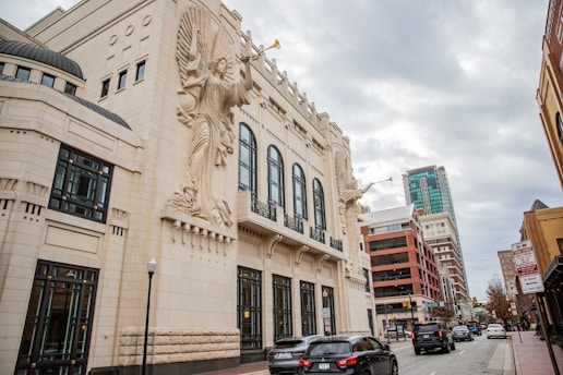 An ornate building with large sculptural figures playing trumpets is prominently featured. The structure has a classical architectural style with tall arched windows. The street in front contains several cars and is lined with other modern buildings, indicating an urban setting. The sky is overcast, adding a muted tone to the scene.