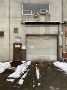 A weathered industrial building features a large, rolled-down metal garage door next to a smaller, rusted brown door with official signage. Above the garage door, there is a rectangular window with an air conditioning unit installed. Snow patches cover the ground in front, creating a stark contrast against the dark, wet pavement.