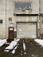 A weathered industrial building features a large, rolled-down metal garage door next to a smaller, rusted brown door with official signage. Above the garage door, there is a rectangular window with an air conditioning unit installed. Snow patches cover the ground in front, creating a stark contrast against the dark, wet pavement.