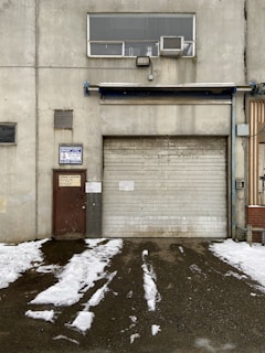 A weathered industrial building features a large, rolled-down metal garage door next to a smaller, rusted brown door with official signage. Above the garage door, there is a rectangular window with an air conditioning unit installed. Snow patches cover the ground in front, creating a stark contrast against the dark, wet pavement.