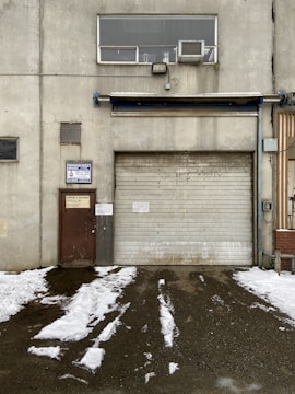 A weathered industrial building features a large, rolled-down metal garage door next to a smaller, rusted brown door with official signage. Above the garage door, there is a rectangular window with an air conditioning unit installed. Snow patches cover the ground in front, creating a stark contrast against the dark, wet pavement.