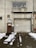 A weathered industrial building features a large, rolled-down metal garage door next to a smaller, rusted brown door with official signage. Above the garage door, there is a rectangular window with an air conditioning unit installed. Snow patches cover the ground in front, creating a stark contrast against the dark, wet pavement.
