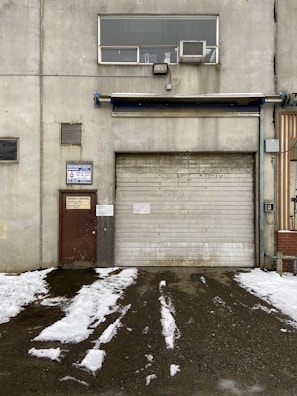 A weathered industrial building features a large, rolled-down metal garage door next to a smaller, rusted brown door with official signage. Above the garage door, there is a rectangular window with an air conditioning unit installed. Snow patches cover the ground in front, creating a stark contrast against the dark, wet pavement.