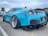 A bright blue sports car with wide tires and custom body kit is parked on a concrete surface. The rear of the car features dual exhaust pipes and a large spoiler. There is a palm tree in the background and another car partially visible on the left side.