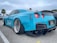 A bright blue sports car with wide tires and custom body kit is parked on a concrete surface. The rear of the car features dual exhaust pipes and a large spoiler. There is a palm tree in the background and another car partially visible on the left side.