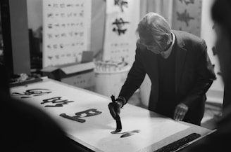 An elderly person is engaged in calligraphy, focused on writing with a brush on a large sheet of paper. The background contains several calligraphy artworks and scrolls, indicating a studio or artistic setting. The image is in black and white, adding a timeless and classic feel.