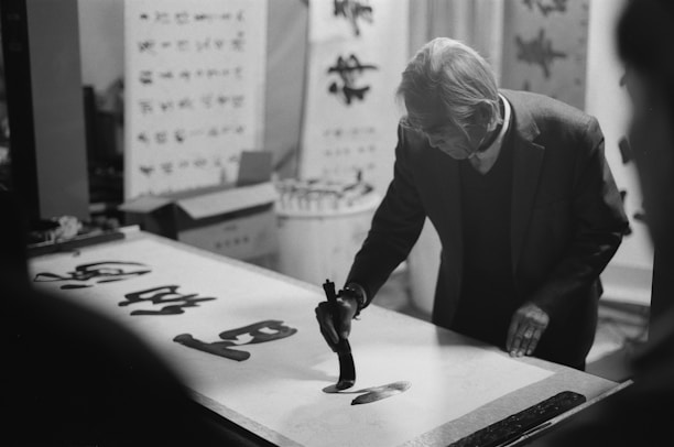 An elderly person is engaged in calligraphy, focused on writing with a brush on a large sheet of paper. The background contains several calligraphy artworks and scrolls, indicating a studio or artistic setting. The image is in black and white, adding a timeless and classic feel.
