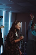 A woman holding a microphone is singing passionately. Another person nearby is raising their hand, possibly enjoying the music. The setting appears to be indoors, with bright vertical lights in the background.