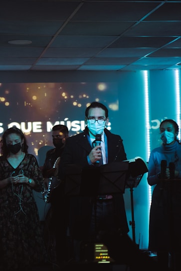 A group of people standing in a dimly lit room with a stage-like setting. They are wearing face masks and appear to be engaged in a musical or speaking event. The person in the center is holding a microphone and a book, suggesting they might be leading the event. The background features a screen with faint lights and text.