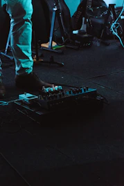 Close-up of a guitarist's hands adjusting wireless gear on a pedalboard in a dimly lit rehearsal space.