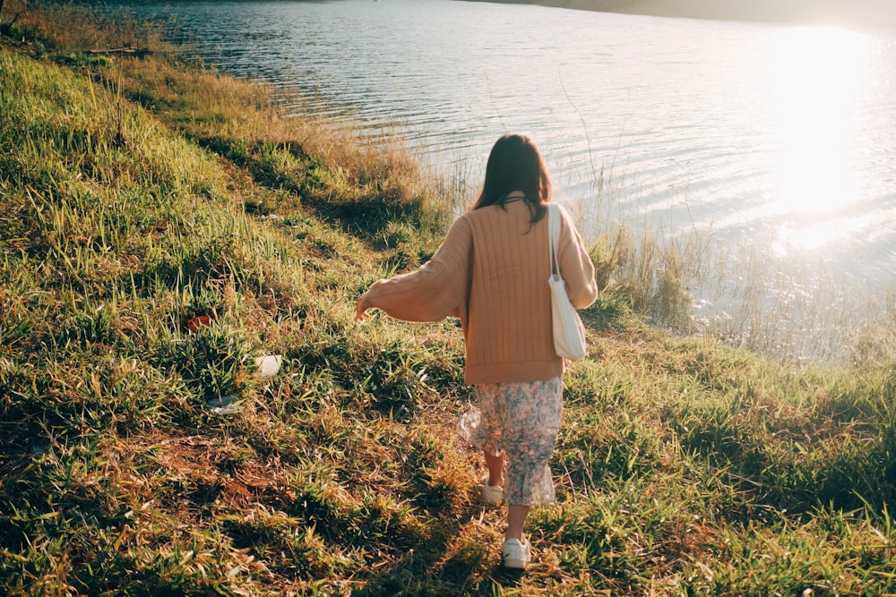 a woman walking along a grass covered hillside next to a body of water