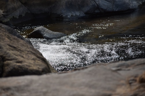 Sunlight sparkles on gently flowing water, surrounded by large rocks. The scene is serene and natural, with a subtle contrast between the dark hues of the rocks and the shimmering surface of the water.