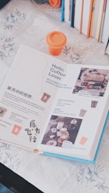 An open book with pages dedicated to coffee, displaying text, images of beverages, and accompanying graphics. An orange cup with a lid is situated above the book. Several books with colorful spines are arranged vertically in the background on the right side. The book is on a floral-patterned tablecloth.
