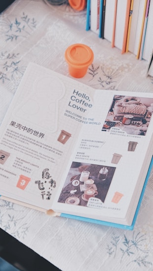 An open book with pages dedicated to coffee, displaying text, images of beverages, and accompanying graphics. An orange cup with a lid is situated above the book. Several books with colorful spines are arranged vertically in the background on the right side. The book is on a floral-patterned tablecloth.
