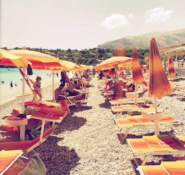 A pebble beach lined with orange and yellow sun umbrellas and lounge chairs. People are sunbathing and swimming in the clear blue water. A backdrop of green hills and a sunny sky enhances the vibrant atmosphere.