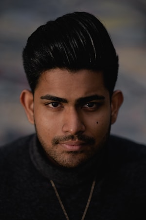 A close-up portrait of a person with a well-groomed beard and styled hair, wearing a dark turtleneck sweater. The background is blurred with neutral tones, emphasizing the subject's features.